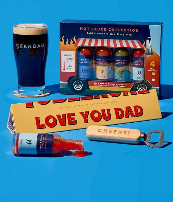 Father’s Day gift arrangement on a blue background featuring a Grandad pint glass, hot sauce gift set, Toblerone bar with Love You Dad message, bottle opener, and spilled sauce bottle